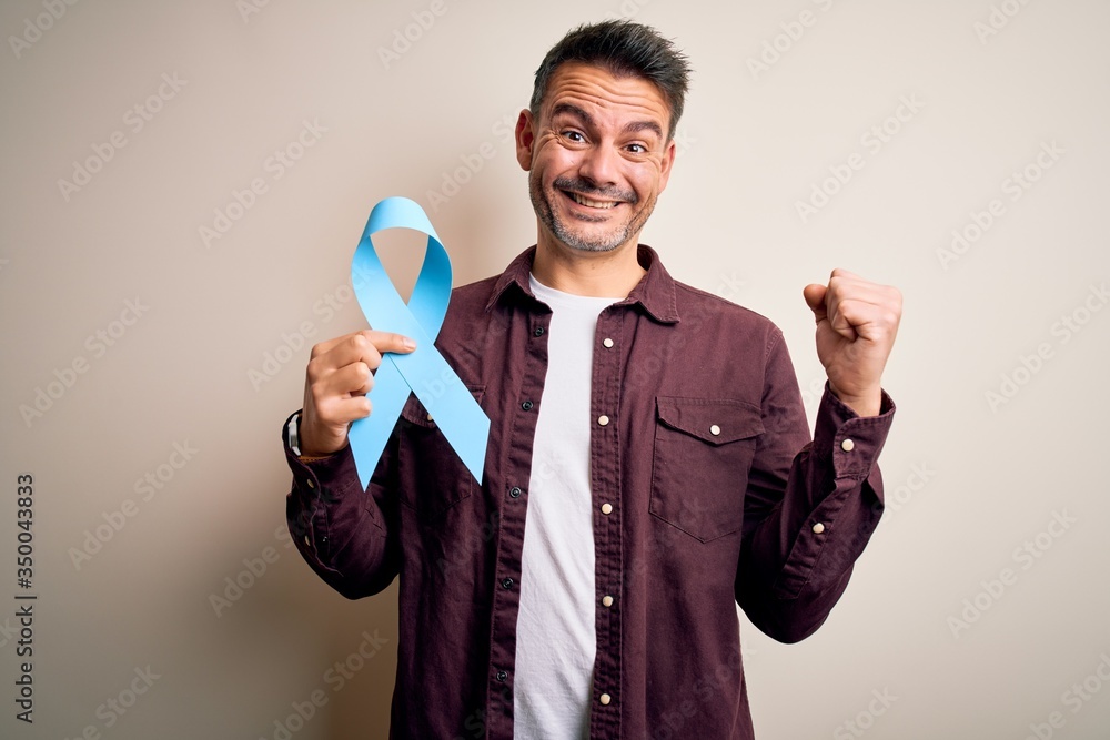 Young handsome man holding blue cancer ribbon over isolated white background screaming proud and celebrating victory and success very excited, cheering emotion Young handsome man holding blue cancer ribbon over isolated white background screaming proud and celebrating victory and success very excited, cheering emotion