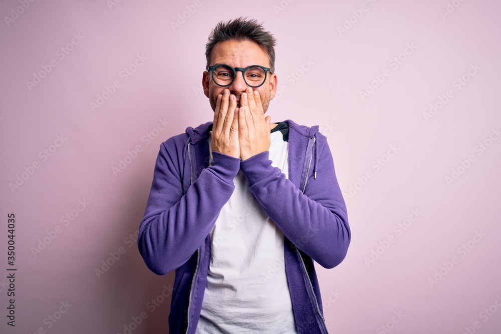 Young handsome man wearing purple sweatshirt and glasses standing over pink background laughing and embarrassed giggle covering mouth with hands, gossip and scandal concept Young handsome man wearing purple sweatshirt and glasses standing over pink background laughing and embarrassed giggle covering mouth with hands, gossip and scandal concept