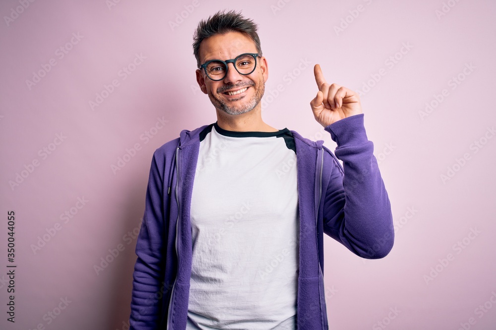 Young handsome man wearing purple sweatshirt and glasses standing over pink background showing and pointing up with finger number one while smiling confident and happy. Young handsome man wearing purple sweatshirt and glasses standing over pink background showing and pointing up with finger number one while smiling confident and happy.