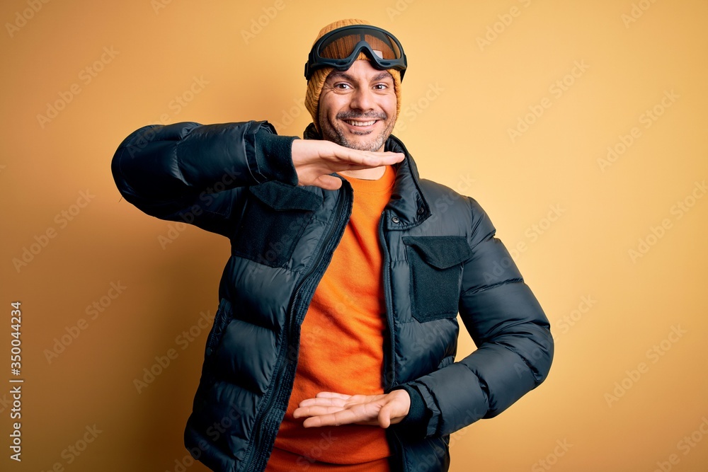 Young handsome skier man skiing wearing snow sportswear using ski goggles gesturing with hands showing big and large size sign, measure symbol. Smiling looking at the camera. Measuring concept.