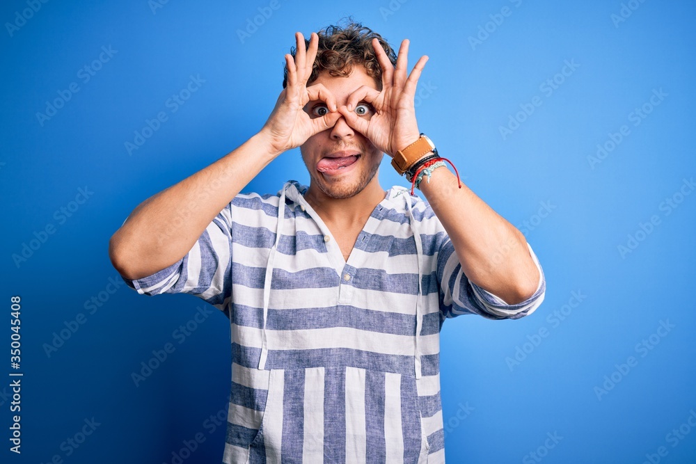 Young blond handsome man with curly hair wearing casual striped sweatshirt doing ok gesture like binoculars sticking tongue out, eyes looking through fingers. Crazy expression. Young blond handsome man with curly hair wearing casual striped sweatshirt doing ok gesture like binoculars sticking tongue out, eyes looking through fingers. Crazy expression.