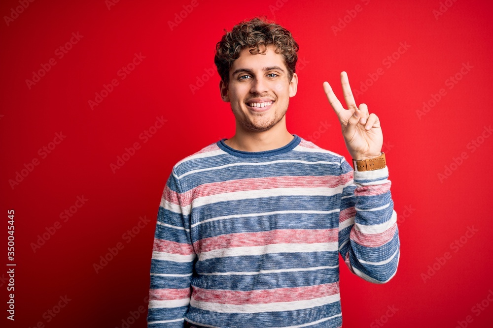 Young blond handsome man with curly hair wearing striped sweater over red background smiling with happy face winking at the camera doing victory sign. Number two. Young blond handsome man with curly hair wearing striped sweater over red background smiling with happy face winking at the camera doing victory sign. Number two.