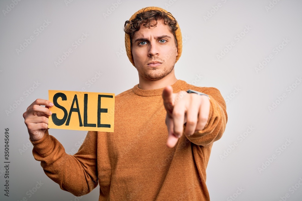 Young blond man with curly hair holding sale poster standing over isolated white background pointing with finger to the camera and to you, hand sign, positive and confident gesture from the front Young blond man with curly hair holding sale poster standing over isolated white background pointing with finger to the camera and to you, hand sign, positive and confident gesture from the front
