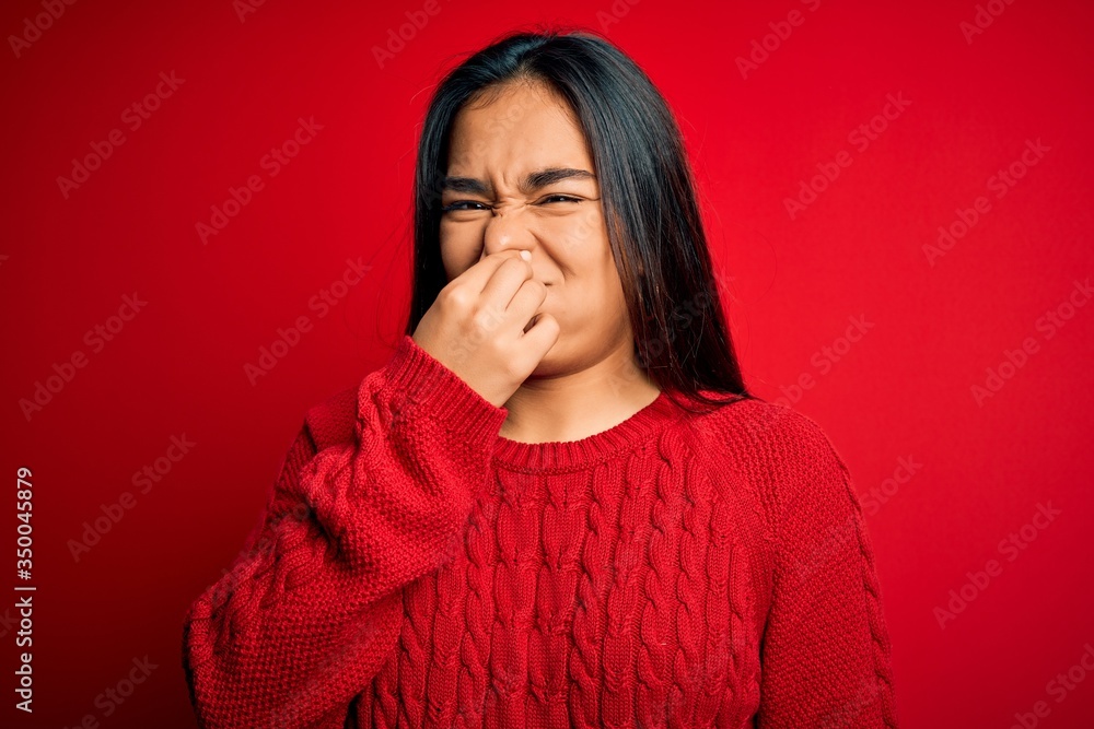 Young beautiful asian woman wearing casual sweater standing over isolated red background smelling something stinky and disgusting, intolerable smell, holding breath with fingers on nose. Bad smell Young beautiful asian woman wearing casual sweater standing over isolated red background smelling something stinky and disgusting, intolerable smell, holding breath with fingers on nose. Bad smell