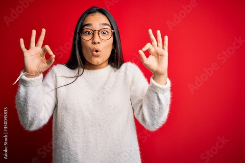 Young beautiful asian woman wearing casual sweater and glasses over red background looking surprised and shocked doing ok approval symbol with fingers. Crazy expression © Krakenimages.com