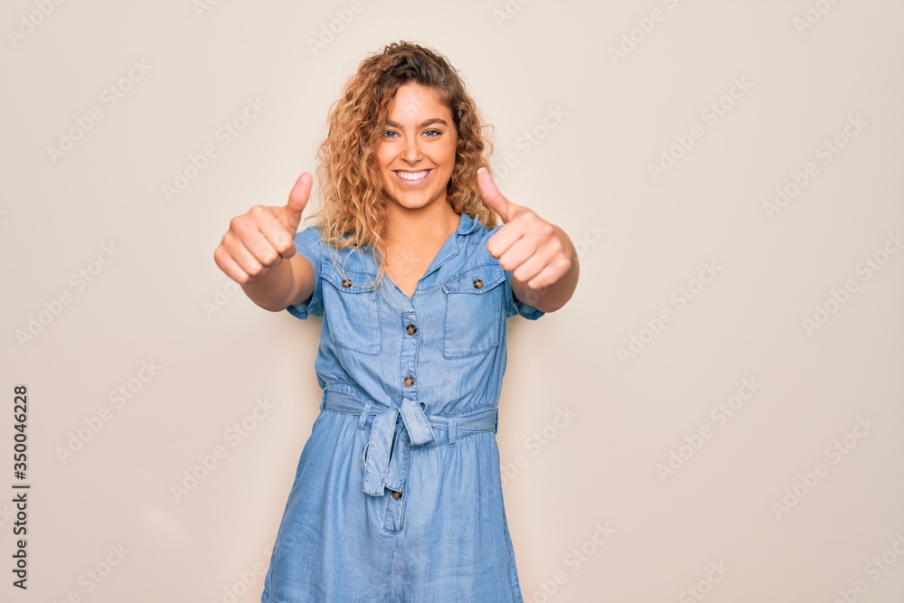 Young beautiful woman with blue eyes wearing casual denim dress over white background approving doing positive gesture with hand, thumbs up smiling and happy for success. Winner gesture. Young beautiful woman with blue eyes wearing casual denim dress over white background approving doing positive gesture with hand, thumbs up smiling and happy for success. Winner gesture.