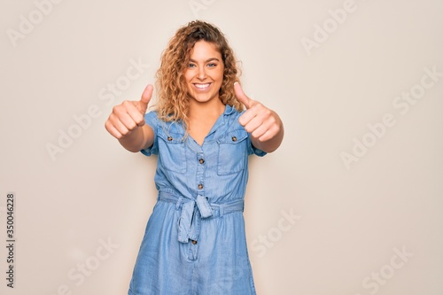 Young beautiful woman with blue eyes wearing casual denim dress over white background approving doing positive gesture with hand, thumbs up smiling and happy for success. Winner gesture.