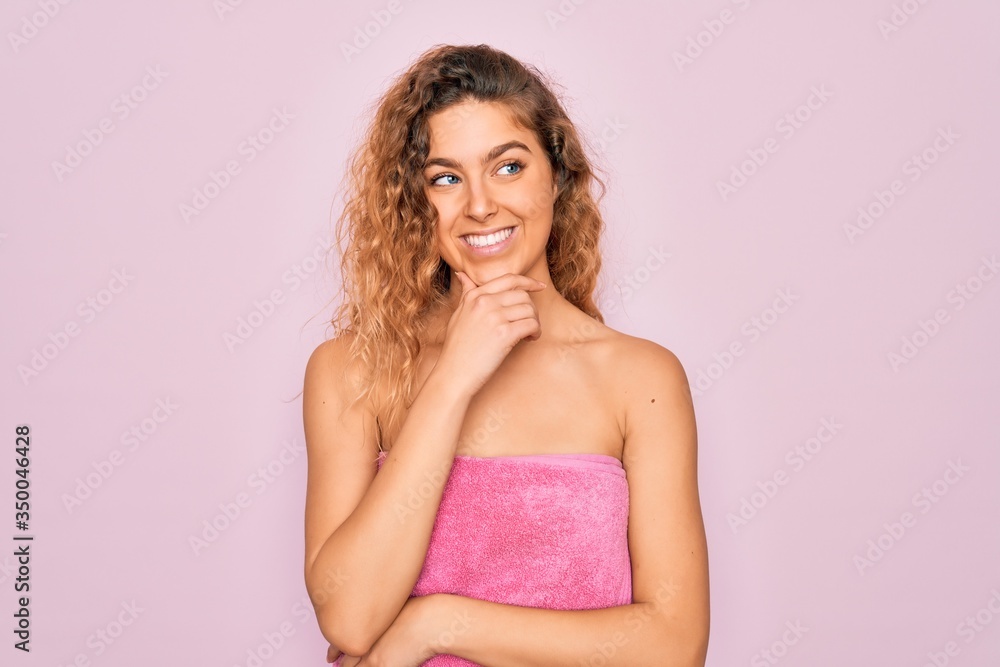 Beautiful blonde woman with blue eyes wearing towel shower after bath over pink background with hand on chin thinking about question, pensive expression. Smiling with thoughtful face. Doubt concept. Beautiful blonde woman with blue eyes wearing towel shower after bath over pink background with hand on chin thinking about question, pensive expression. Smiling with thoughtful face. Doubt concept.