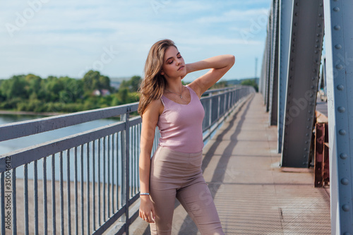 Fototapeta Naklejka Na Ścianę i Meble -  Pretty young woman posing on the old rusty transport bridge over the river during sunset.
