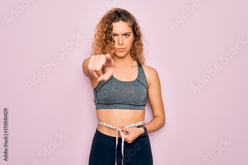 Young blonde sporty woman with blue eyes controlling weight using tape measure on waist pointing with finger to the camera and to you, hand sign, positive and confident gesture from the front © Krakenimages.com