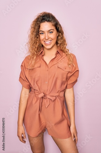 Young beautiful blonde woman with curly hair smiling happy and confident. Standing with smile on face wearing casual dress over isolated pink background