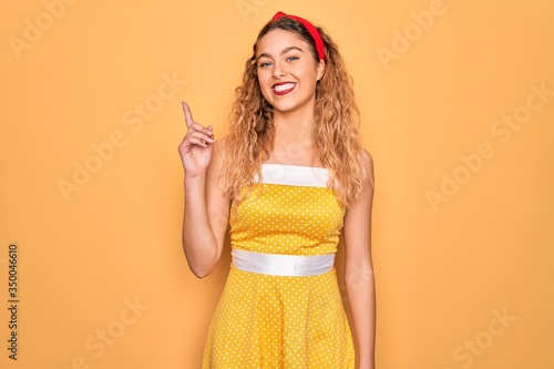 Beautiful blonde pin-up woman with blue eyes wearing diadem standing over yellow background showing and pointing up with finger number one while smiling confident and happy.