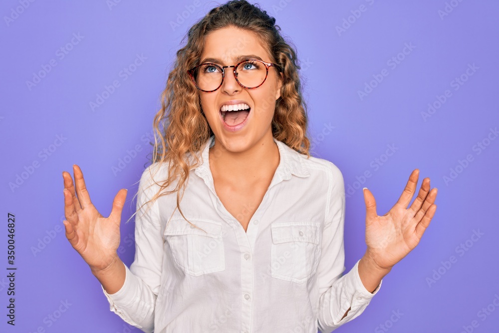Young beautiful woman with blue eyes wearing casual shirt and glasses over purple background crazy and mad shouting and yelling with aggressive expression and arms raised. Frustration concept. Young beautiful woman with blue eyes wearing casual shirt and glasses over purple background crazy and mad shouting and yelling with aggressive expression and arms raised. Frustration concept.