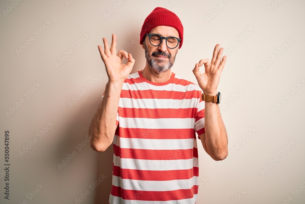 Naklejka premium Middle age hoary man wearing striped t-shirt glasses and cap over isolated white background relax and smiling with eyes closed doing meditation gesture with fingers. Yoga concept.
