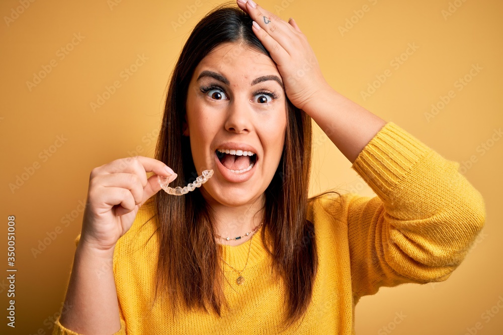 Young beautiful woman holding dental orthodontic aligner corrector over yellow background stressed with hand on head, shocked with shame and surprise face, angry and frustrated. Fear and upset Young beautiful woman holding dental orthodontic aligner corrector over yellow background stressed with hand on head, shocked with shame and surprise face, angry and frustrated. Fear and upset