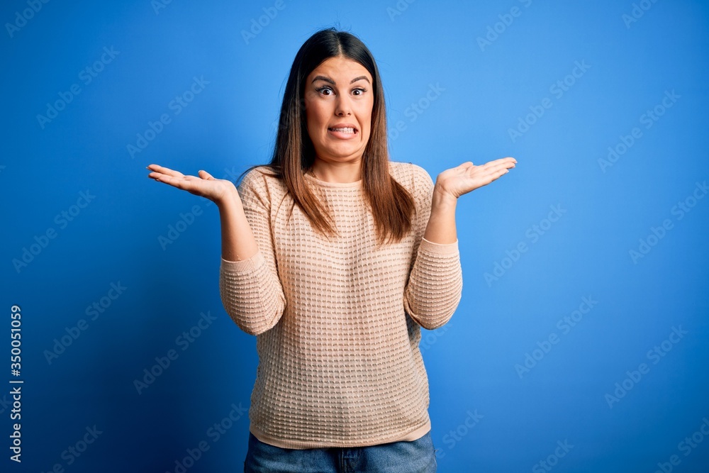 Young beautiful woman wearing casual sweater over blue background clueless and confused expression with arms and hands raised. Doubt concept. Young beautiful woman wearing casual sweater over blue background clueless and confused expression with arms and hands raised. Doubt concept.