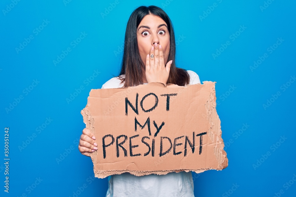Young beautiful woman on disagreement holding banner with not my president message covering mouth with hand, shocked and afraid for mistake. Surprised expression Young beautiful woman on disagreement holding banner with not my president message covering mouth with hand, shocked and afraid for mistake. Surprised expression
