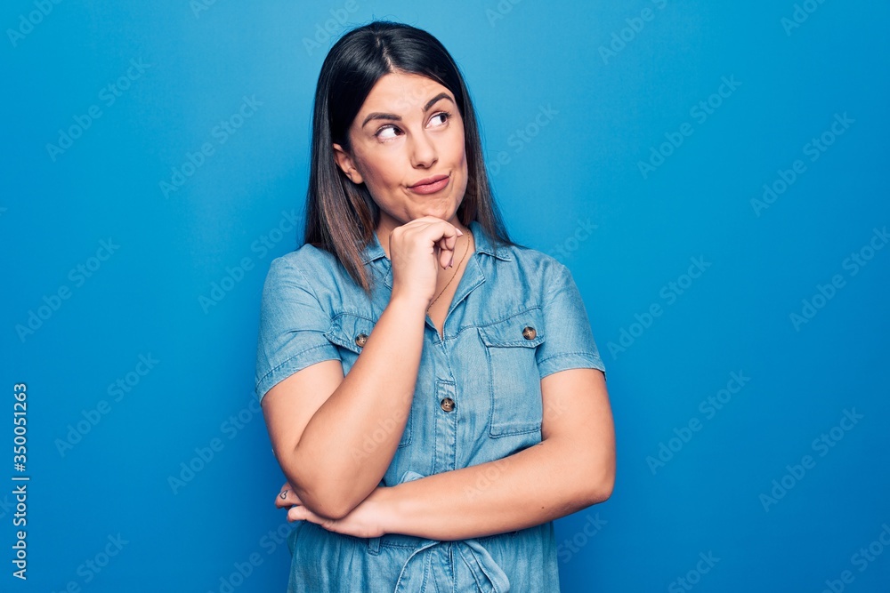Young beautiful brunette woman wearing casual denim dress over isolated blue background thinking concentrated about doubt with finger on chin and looking up wondering Young beautiful brunette woman wearing casual denim dress over isolated blue background thinking concentrated about doubt with finger on chin and looking up wondering