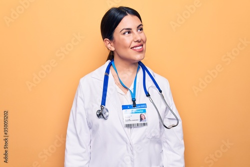Young beautiful doctor woman wearing stethoscope and id card over isolated yellow background looking to side  relax profile pose with natural face and confident smile.