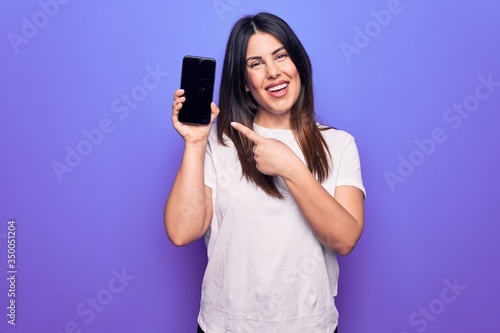 Young beautiful brunette woman holding smartphone showing screen over purple background smiling happy pointing with hand and finger