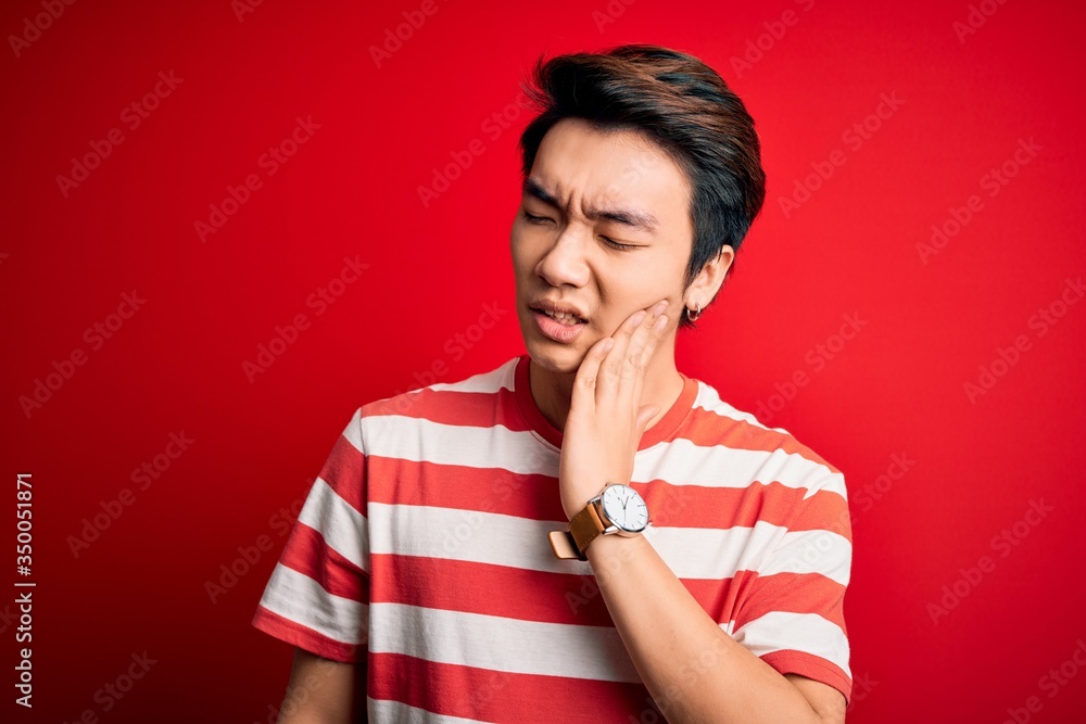 Young handsome chinese man wearing casual striped t-shirt standing over red background touching mouth with hand with painful expression because of toothache or dental illness on teeth. Dentist Young handsome chinese man wearing casual striped t-shirt standing over red background touching mouth with hand with painful expression because of toothache or dental illness on teeth. Dentist