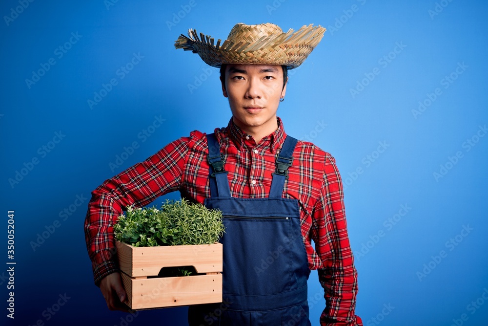 Young handsome chinese farmer man wearing apron and straw hat holding box with plants with a confident expression on smart face thinking serious