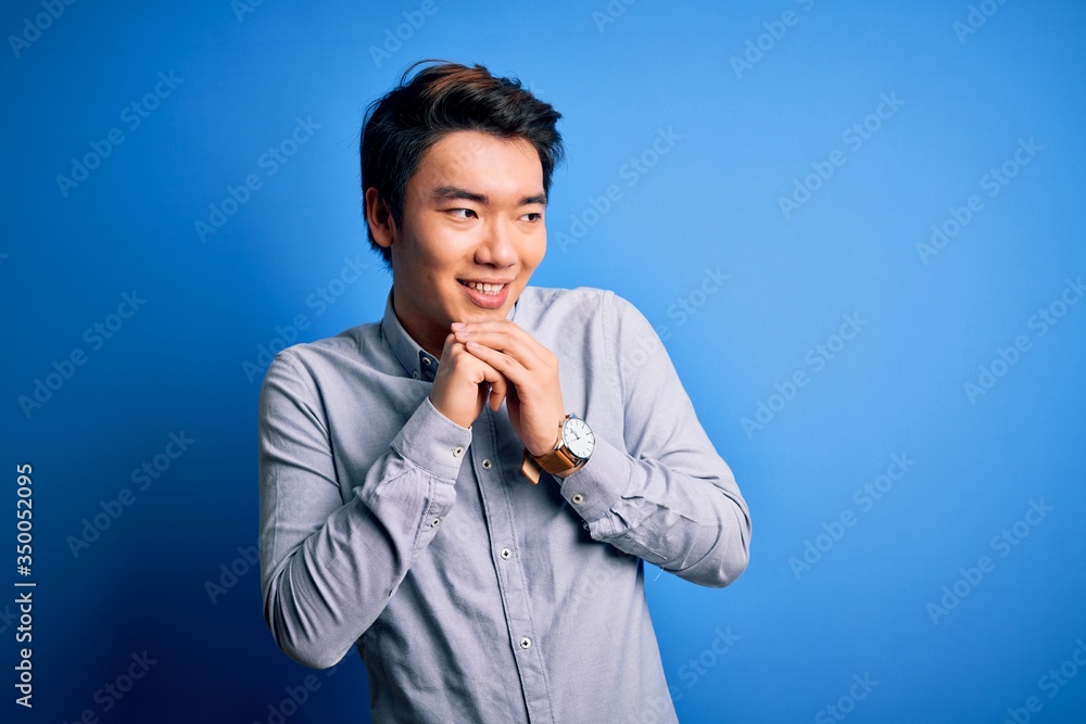 Young handsome chinese man wearing casual shirt standing over isolated blue background laughing nervous and excited with hands on chin looking to the side Young handsome chinese man wearing casual shirt standing over isolated blue background laughing nervous and excited with hands on chin looking to the side