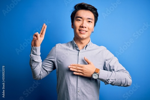 Young handsome chinese man wearing casual shirt standing over isolated blue background smiling swearing with hand on chest and fingers up, making a loyalty promise oath
