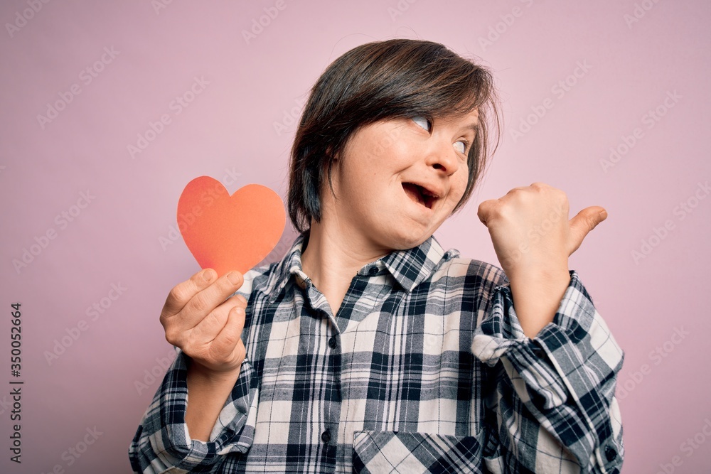 Young down syndrome romantic woman holding red heart paper shape over pink background pointing and showing with thumb up to the side with happy face smiling Young down syndrome romantic woman holding red heart paper shape over pink background pointing and showing with thumb up to the side with happy face smiling