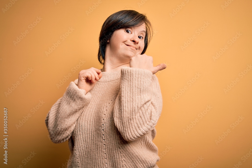 Young down syndrome woman wearing casual sweater over yellow background smiling with happy face looking and pointing to the side with thumb up. Young down syndrome woman wearing casual sweater over yellow background smiling with happy face looking and pointing to the side with thumb up.