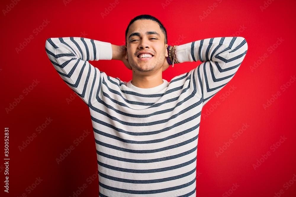 Young brazilian man wearing casual striped t-shirt standing over isolated red background relaxing and stretching, arms and hands behind head and neck smiling happy Young brazilian man wearing casual striped t-shirt standing over isolated red background relaxing and stretching, arms and hands behind head and neck smiling happy