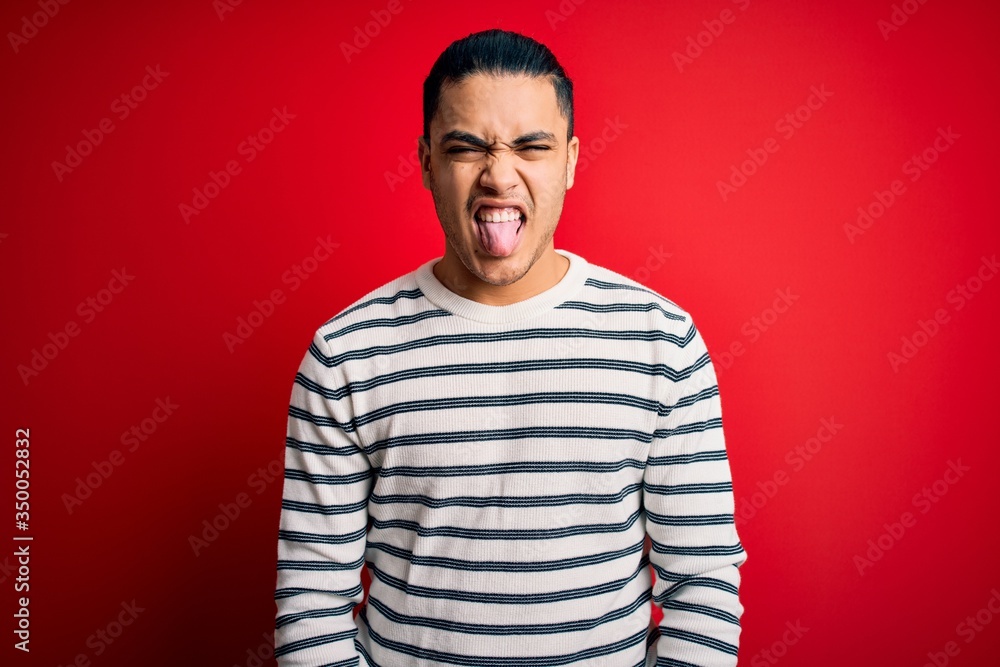 Young brazilian man wearing casual striped t-shirt standing over isolated red background sticking tongue out happy with funny expression. Emotion concept. Young brazilian man wearing casual striped t-shirt standing over isolated red background sticking tongue out happy with funny expression. Emotion concept.