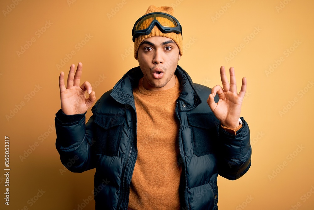 Young brazilian skier man wearing snow sportswear and ski goggles over yellow background looking surprised and shocked doing ok approval symbol with fingers. Crazy expression Young brazilian skier man wearing snow sportswear and ski goggles over yellow background looking surprised and shocked doing ok approval symbol with fingers. Crazy expression
