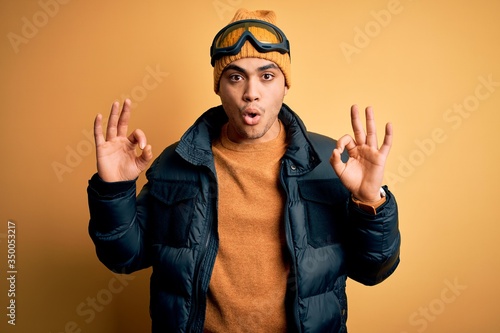 Young brazilian skier man wearing snow sportswear and ski goggles over yellow background looking surprised and shocked doing ok approval symbol with fingers. Crazy expression