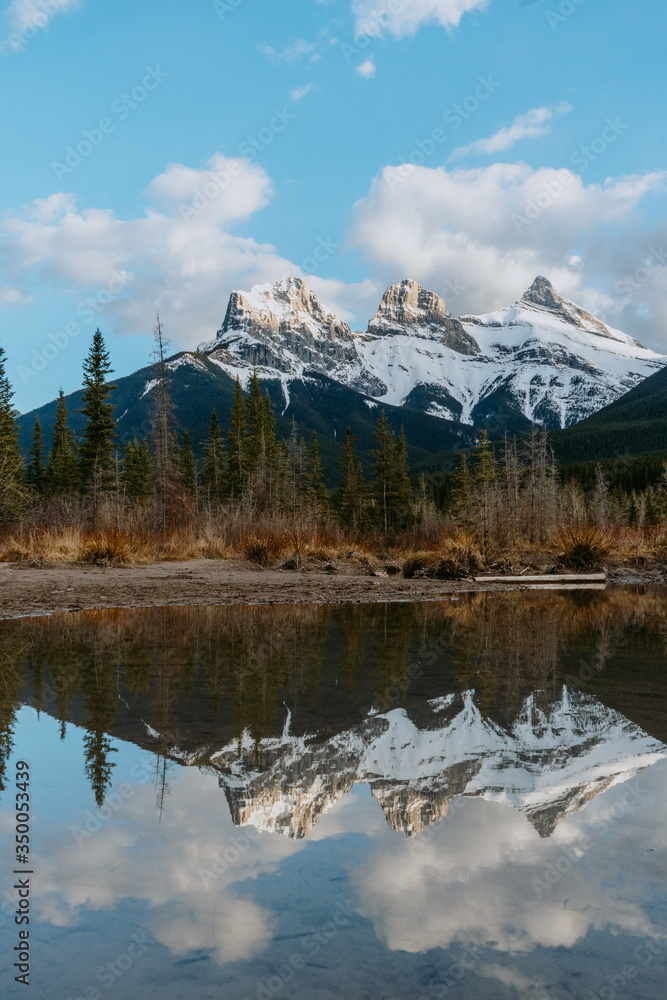 Breathtaking view of the iconic Three Sisters mountain peaks reflection in Policeman's Creek calm water while sunset. Canmore, Alberta, Canada.