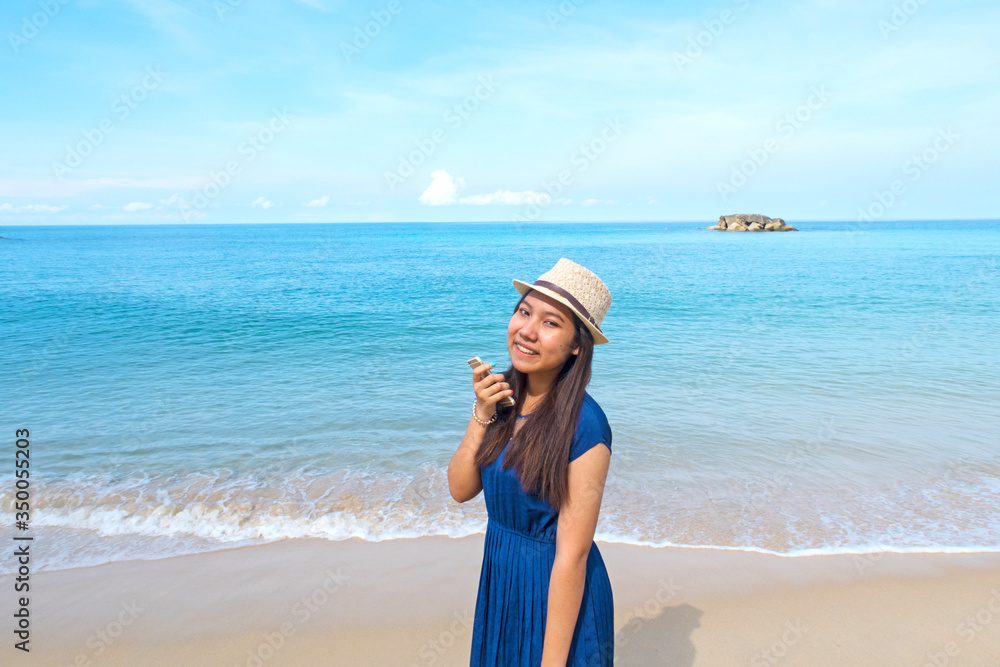 Asian young teenager girl wearing sun hat & cute navy blue dress holding mobile phone or smartphone enjoying & admiring of deep blue sea view with soft calm waves touching tropical summer sand beach Asian young teenager girl wearing sun hat & cute navy blue dress holding mobile phone or smartphone enjoying & admiring of deep blue sea view with soft calm waves touching tropical summer sand beach
