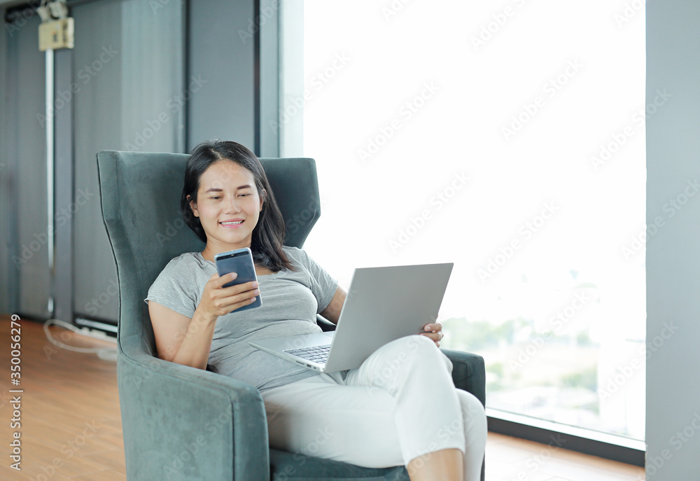 Young beautiful asian woman using smartphone and laptop computer working online at home and feel relaxing on armchair. People during the introvert time at home from the epidemic in thailand. Young beautiful asian woman using smartphone and laptop computer working online at home and feel relaxing on armchair. People during the introvert time at home from the epidemic in thailand.