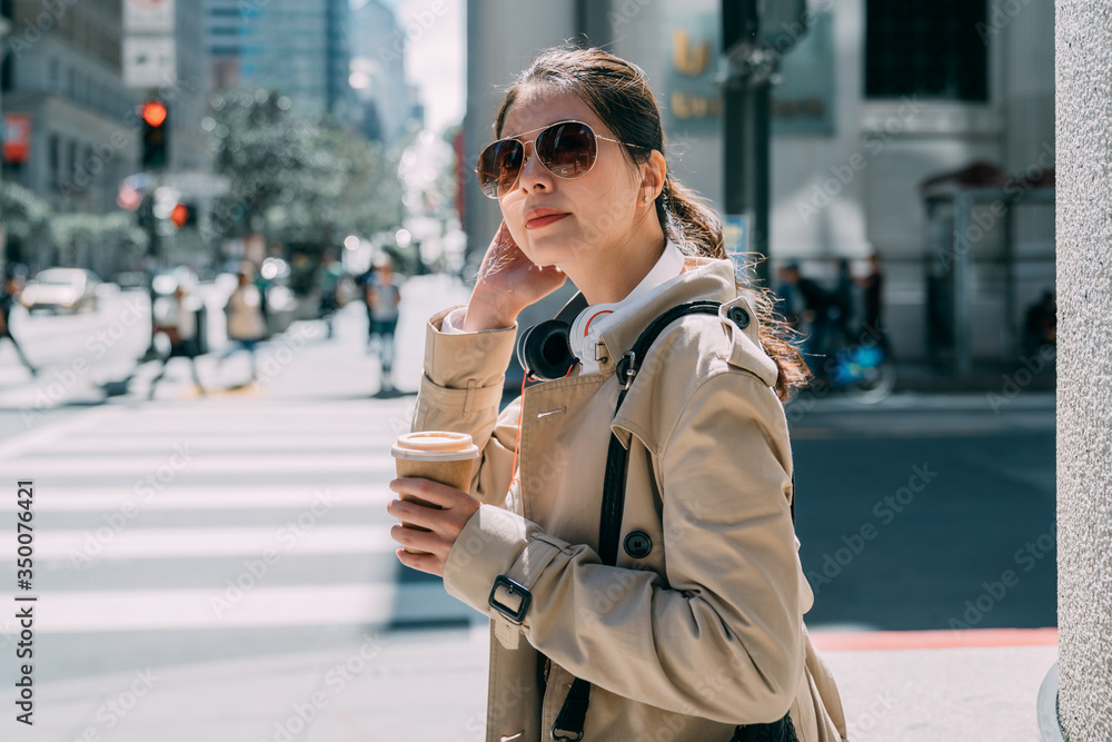 Fototapeta premium Young businesswoman with headphones and to go coffee on street. elegant office lady waiting to cross zebra road in busy urban. female worker with earphones and hands holding paper cup of hot beverage