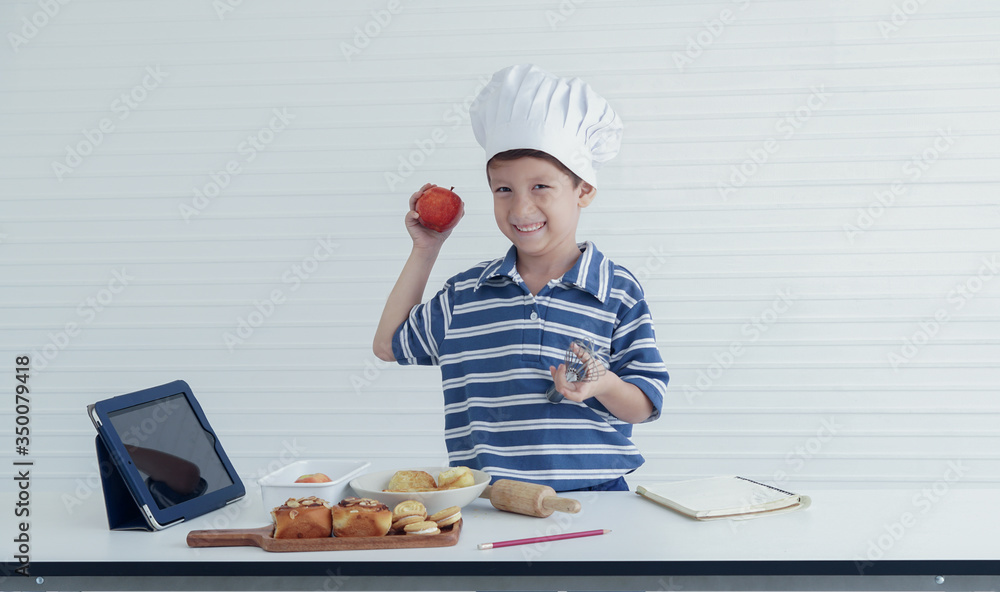 A little Caucasian boy use tablet search how to cook and ingredients to prepare food for cooking in studio kitchen. He holding apple and whisk in his two hands looking at camera with smiley face A little Caucasian boy use tablet search how to cook and ingredients to prepare food for cooking in studio kitchen. He holding apple and whisk in his two hands looking at camera with smiley face