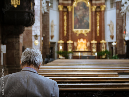Rear view of an elderly man sitting alone in church praying with the ornate altar visible in the distance © raphoto