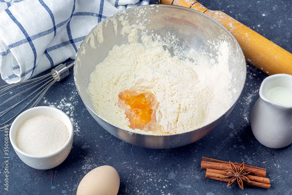 Flour with egg in a metal bowl among ingredients and utensils for cooking cake (flour, egg, milk, sugar, rolling pin, towel) on dark table. The concept of making dough for baking. Close up - obrazy, fototapety, plakaty 