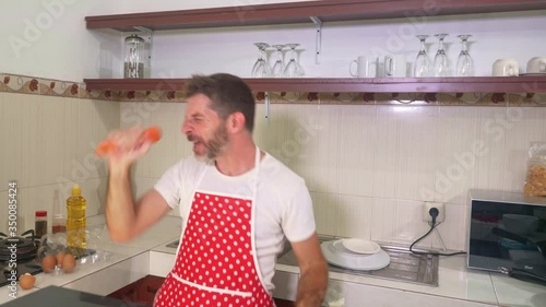 enjoying domestic chores - young cool and attractive man in red apron singing song and dancing crazy happy holding carrot as microphone having fun cooking carefree photo