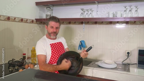 enjoying domestic chores - young cool and attractive man in red apron and washing glove singing song and dancing crazy happy holding pan as if playing guitar excited and carefree photo