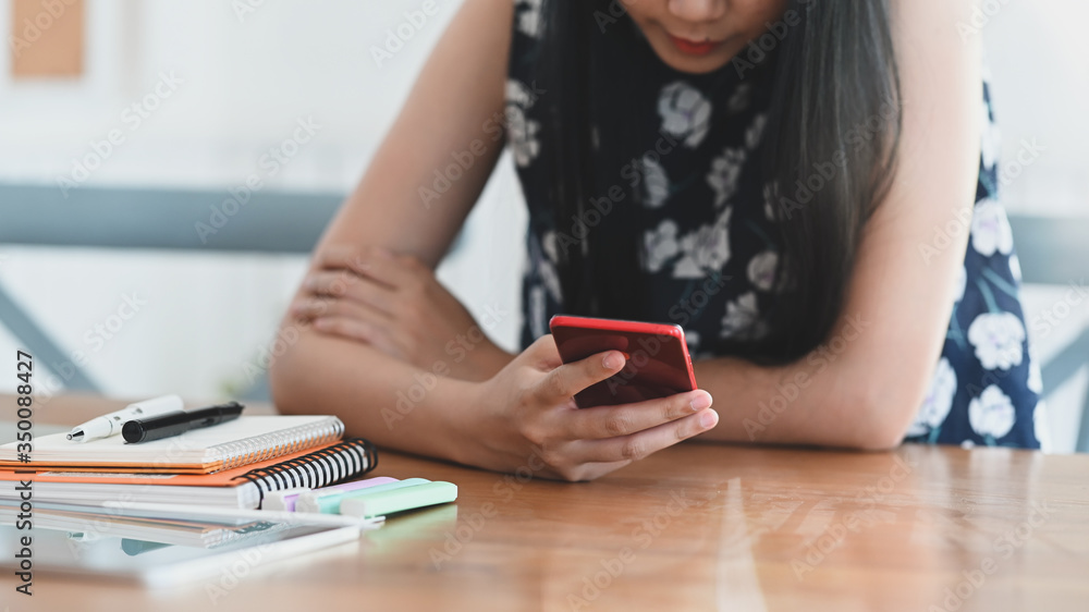 Cropped image of beautiful woman using smartphone while sitting at the wooden working desk that surrounded by office equipment after finished work. Cropped image of beautiful woman using smartphone while sitting at the wooden working desk that surrounded by office equipment after finished work.