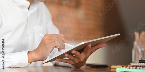 Cropped image of businessman in white shirt drawing on computer tablet in his hand by using a stylus pen while sitting at the wooden working desk over vintage living room as background.