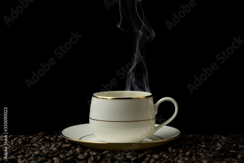 Hot black coffee in white cup with white smoke motion blur up on the air on black background put on coffee beans field