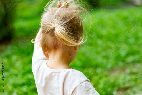 Adorable blond toddler girl in a windy summer day wearing ponytail trying to hold her fluterring hair. Veiw from behind. Copy space