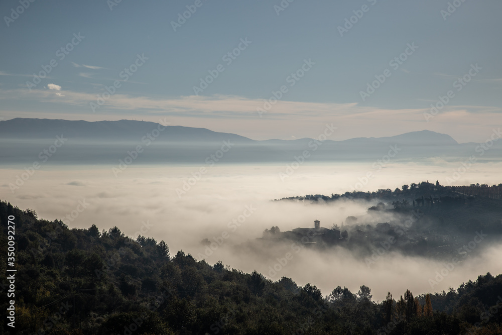 Obraz premium Surreal view of of a little town in Umbria (Italy) almost completely hidden by fog with trees silhouettes in the foreground