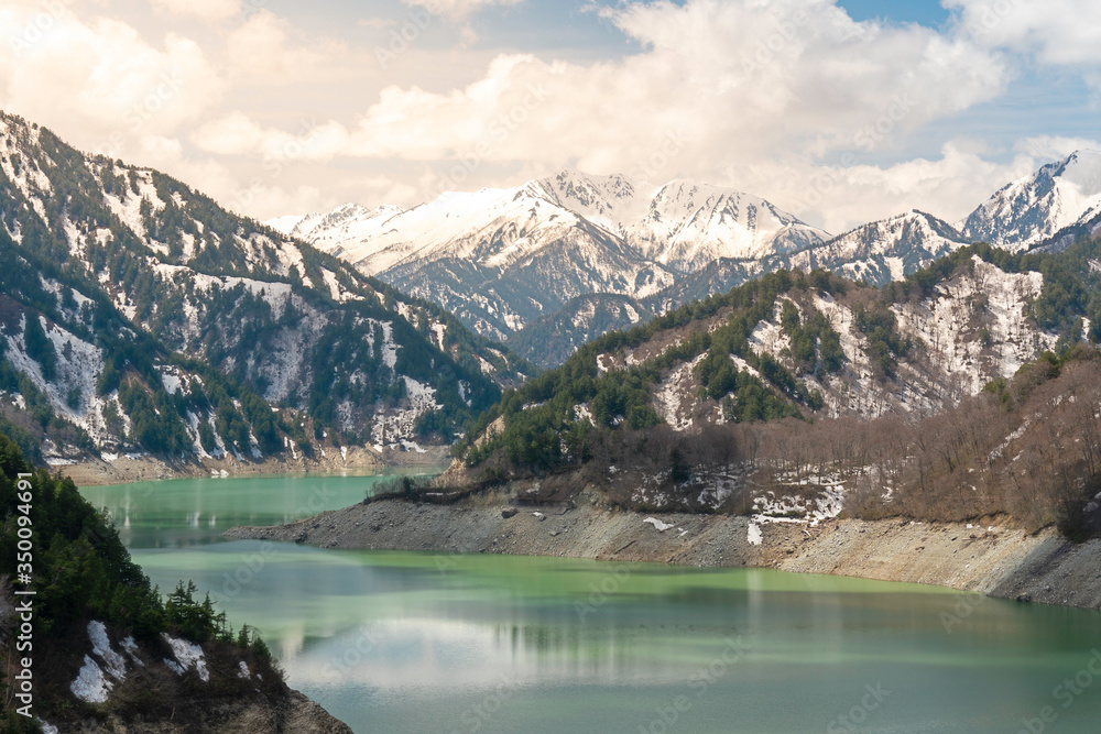 Beautiful nature background and wallpaper of green reservoir lake at Kurobe dam surrounded by snow mountain of Japan Alps under blue sky with cloud and sun light. Top tourist destination in Nagano