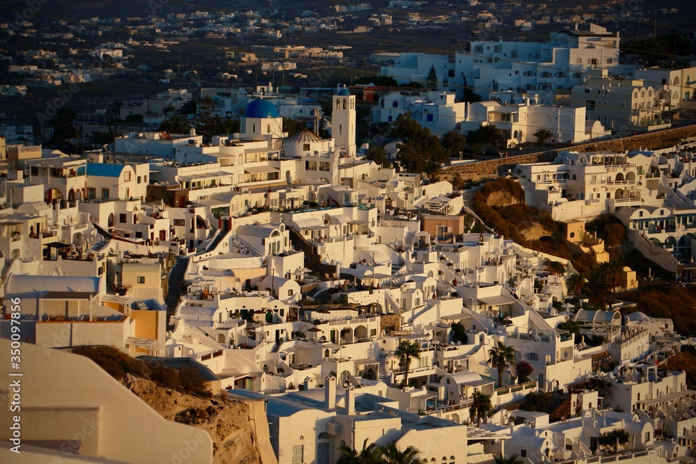 Large side view of Oia city in Santorini (Greece) shot at the golden hour depicting the villas a church and a windmill on the cliff side with their shadow giving a dramatic aspect